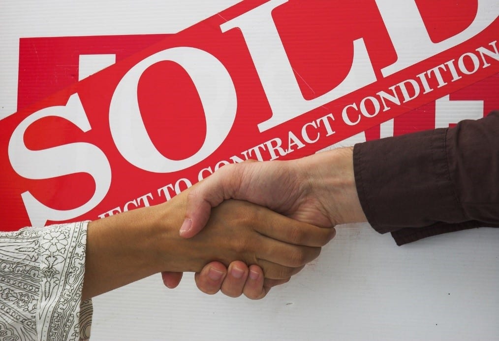 Two hands shaking in front of a sold property sign
